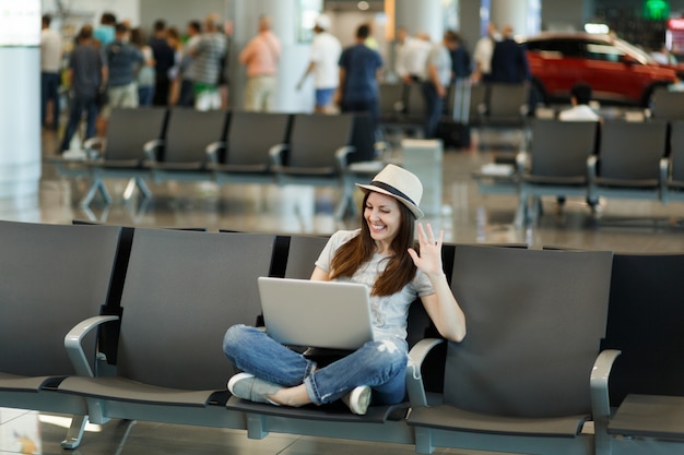 Donna turistica viaggiatrice con cappello che lavora al computer portatile, agitando la mano per salutare in webcam durante la videochiamata, in attesa nella hall dell'aeroporto