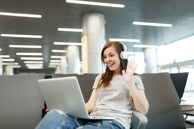 Donna turistica viaggiatrice con cuffie che lavora sulla mano d'onda del laptop per salutare in webcam durante la videochiamata in attesa nella hall dell'aeroporto