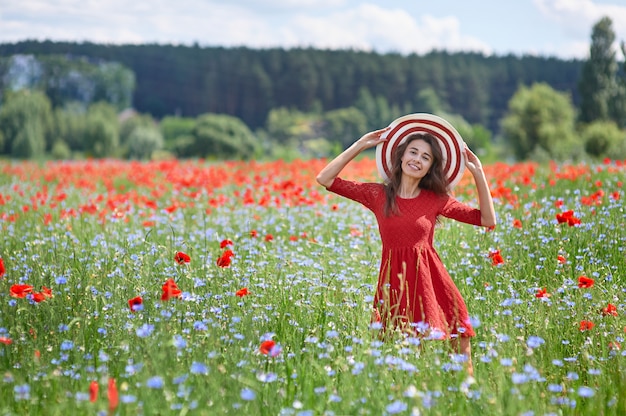 Donna vaga in vestito rosso e un grande cappello a strisce rosso nel campo di papavero fioritura bella erba