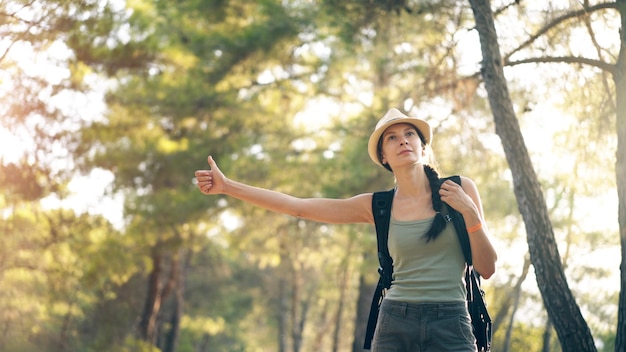 Donna viaggiatore che fa l'autostop su una strada forestale soleggiata Ragazza turistica in cerca di un passaggio per iniziare il suo viaggio