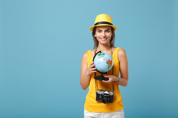 Donna viaggiatrice in abiti casual estivi gialli e cappello con globo e macchina fotografica su blu