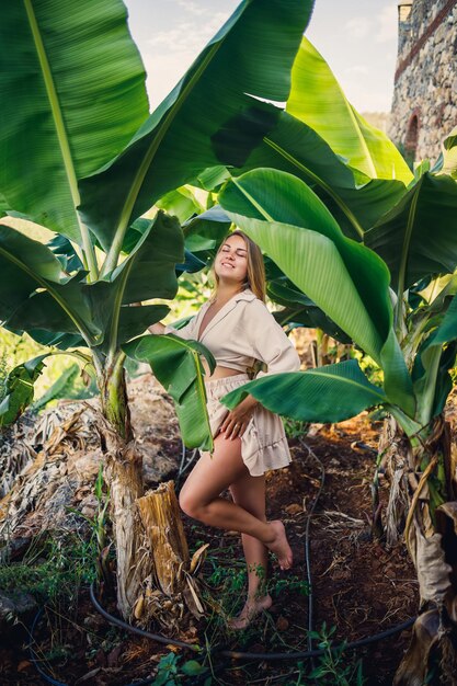 Donna vicino a foglie verdi di cespugli di banane sulla natura in un parco in un luogo tropicale, indossa una gonna beige e una camicetta