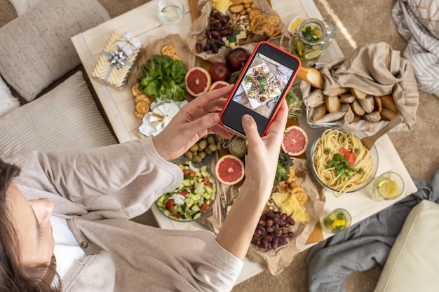Donna vista dall'alto che fotografa la tavola da pranzo con che serve cibo e bevande gustose usa lo smartphone