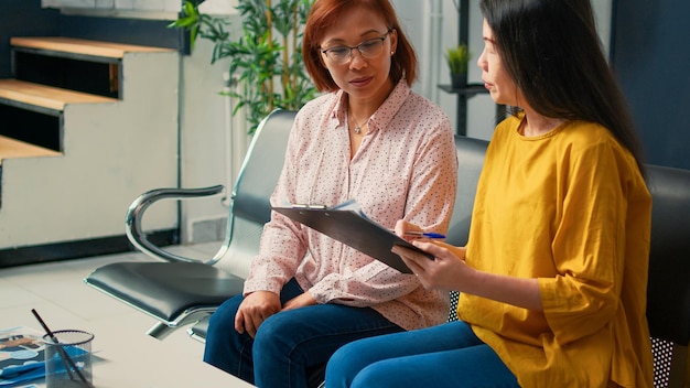 Donne asiatiche che compilano il rapporto di controllo prima della consultazione, aspettano nell'atrio dell'ospedale e parlano di supporto sanitario. Seduto in sala d'attesa per partecipare all'appuntamento medico.