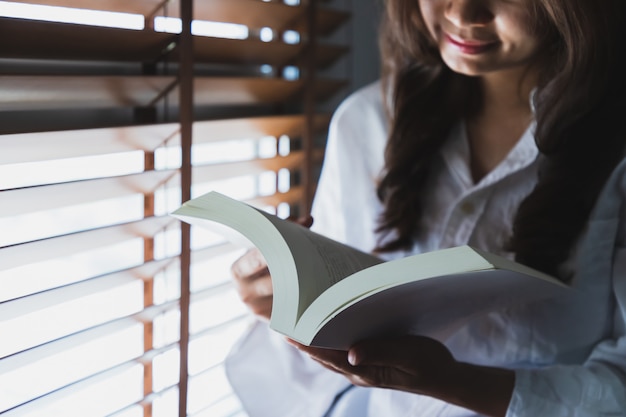 Donne asiatiche che indossano una camicia bianca, leggono un libro bianco vicino alla finestra della camera da letto