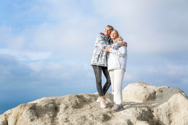 Donne belle in piedi su un'alta montagna sulla costa del mare in una giornata di sole bionde sorridenti che si abbracciano stile di vita attivo e ricreazione altezza piena