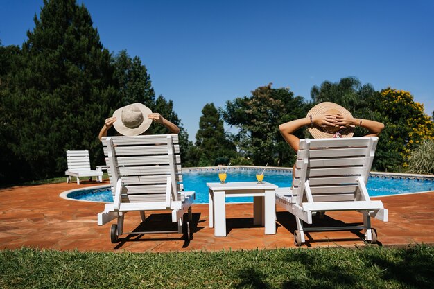 Donne con cappelli sedute sulle sedie a sdraio che prendono il sole vicino alla piscina.