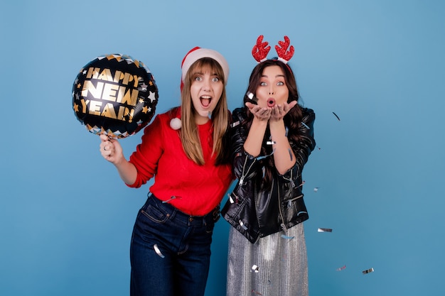 Donne con l'aerostato del buon anno che soffia i coriandoli d'argento che portano il cappello di natale isolato sopra l'azzurro