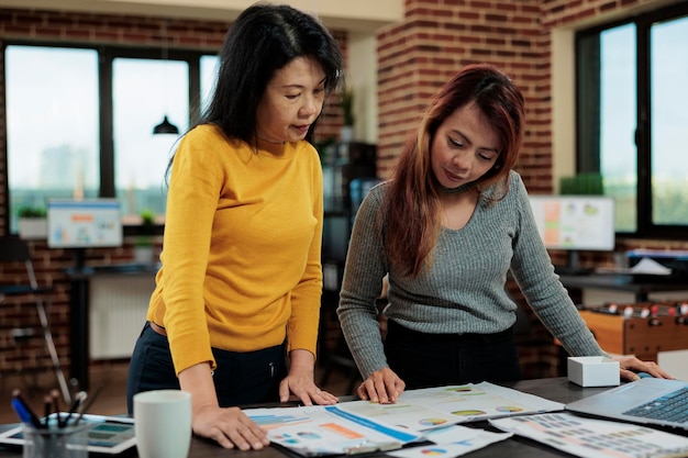 Donne d'affari asiatiche che esaminano le statistiche aziendali che discutono della strategia di gestione mentre lavorano a un progetto aziendale nell'ufficio di avvio. Imprenditrici donne che pianificano un progetto di partenariato