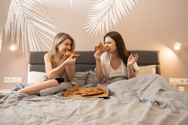 Donne europee felici sdraiate sul letto e mangiando pizza. Diario e scatola di cartone con pizza sul letto. Le amiche allegre si divertono. Concetto di festa delle ragazze a casa. Interno della camera da letto in appartamento moderno