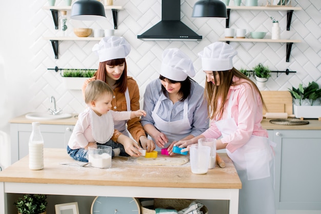 Donne felici in grembiuli bianchi che cuociono insieme, ritagliando le forme dalla pasta del biscotto di zucchero con le formine per biscotti. La piccola neonata aiuta a fare i biscotti insieme a madre, zia e nonna