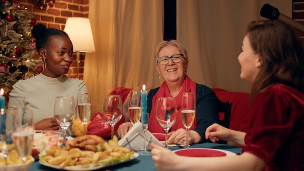 Donne festive felici che si siedono al tavolo della cena di Natale mentre ridono e parlano insieme. Persone gioiose si sono radunate nel soggiorno di casa per celebrare le vacanze tradizionali della stagione degli avvolgitori.