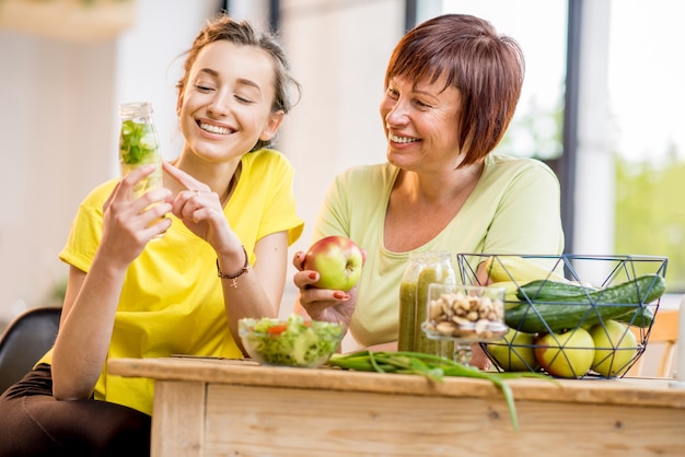 Donne giovani e anziane sedute con cibo sano e bevande fresche dopo l'allenamento sportivo al chiuso sullo sfondo della finestra