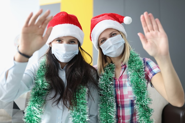 Donne in cappelli rossi di natale e maschere mediche sui loro volti che salutano