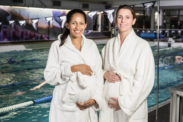 Donne in gravidanza con accappatoio toccando le loro pance in piscina