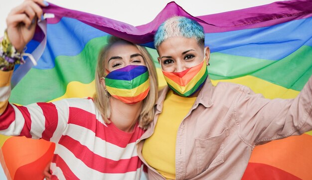 Donne lesbiche felici con bandiera arcobaleno lgbt alla parata dell'orgoglio gay che indossano una maschera colorata di sicurezza per l'epidemia di coronavirus