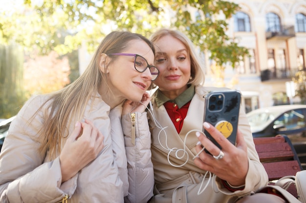 Donne mature adorabili che ascoltano musica insieme sullo smartphone, indossando gli auricolari