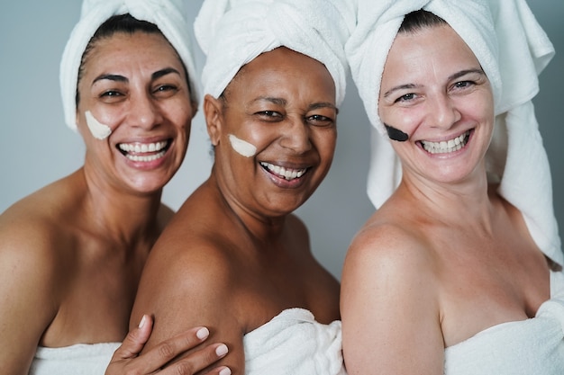 Donne mature che ridono insieme mentre fanno un trattamento di bellezza alla spa usando la maschera per la pelle - Focus principale sul volto femminile africano - Focus principale sulla faccia di casa giusta
