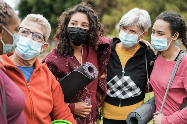 Donne multigenerazionali che si divertono prima della lezione di yoga indossando maschere di sicurezza al parco all'aperto