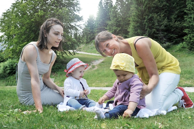 Donne sedute con bambine sull'erba nel parco Due donne amiche che si godono una bella giornata di sole con le loro piccole figlie Concetto di amicizia di maternità e vacanze estive