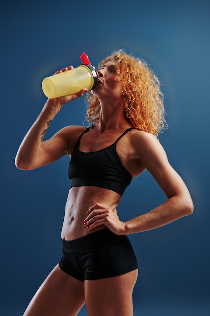 Dopo l'allenamento. Recuperare con l'acqua. Il bodybuilder femminile della testarossa è in studio sullo spazio blu.