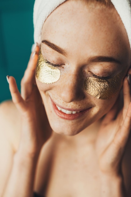 Dopo la doccia donna dai capelli rossi che applica macchie dorate sotto gli occhi toccando il viso con le mani in posa indo