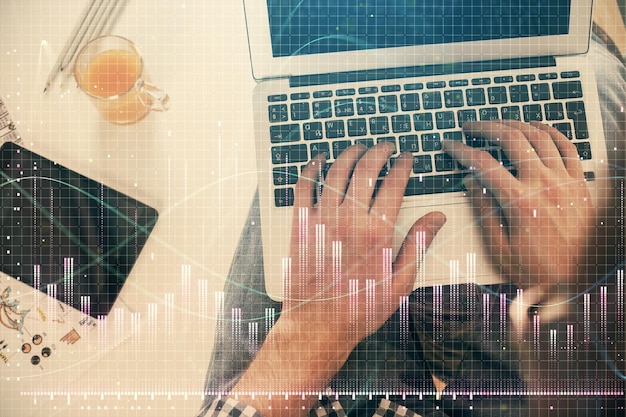 Doppia esposizione delle mani dell'uomo che digitano sulla tastiera del laptop e sul disegno dell'ologramma del grafico forex Vista dall'alto Concetto di mercati finanziari