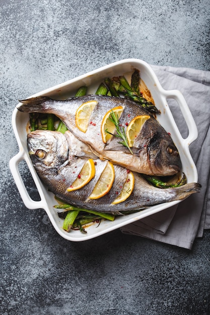 Dorado di pesce al forno con asparagi verdi in teglia da forno in ceramica bianca su fondo di cemento rustico grigio, vista dall'alto. Cena sana con concetto di pesce, dieta e alimentazione pulita