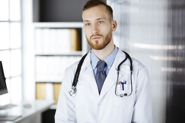 Dottore barbuto rosso in piedi in clinica vicino al suo posto di lavoro. Ritratto di medico. Concept di medicina e assistenza sanitaria.