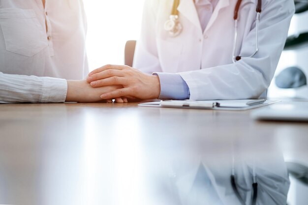 Dottore e paziente seduti l'uno vicino all'altro al tavolo di legno in clinica. Le mani del medico rassicurano la donna. Il concetto di medicina.
