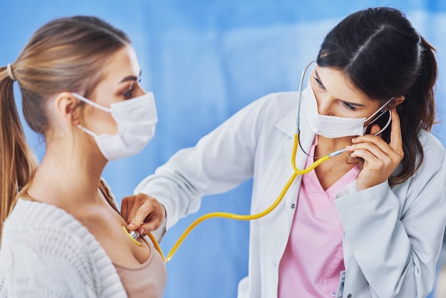 dottore in maschera che controlla una paziente usando lo stetoscopio