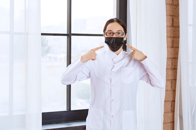 Dottore in maschera nera e occhiali in piedi vicino alla finestra e indicando la sua maschera.