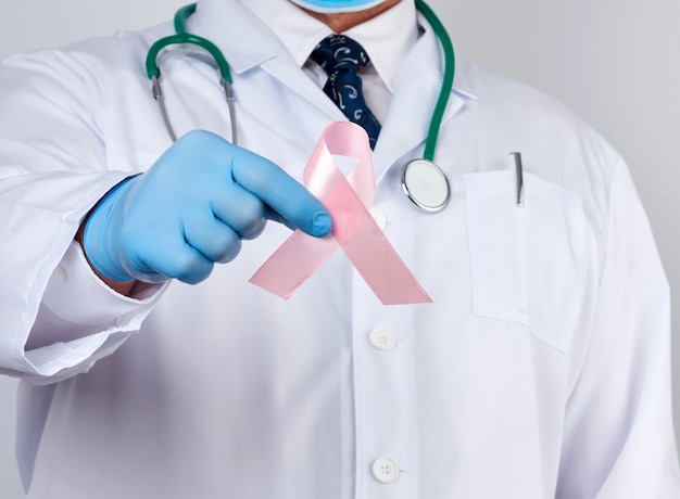 Dottore maschio in camice bianco e cravatta si erge e tiene un nastro di seta rosa a forma di anello, indossando guanti medici sterili blu