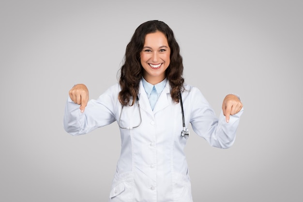 Dottoressa amichevole in camice bianco punta le dita verso il basso nello spazio libero e sorride isolata sul grigio