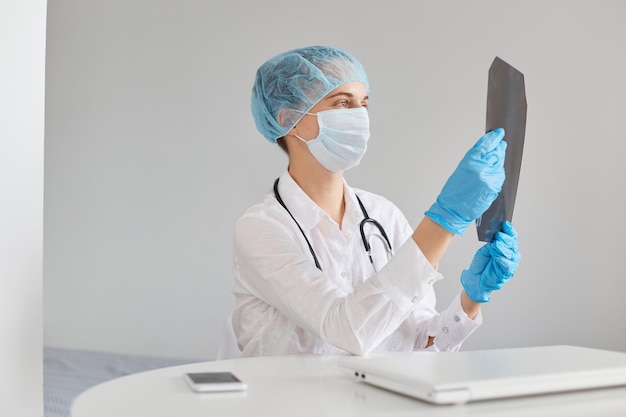 Dottoressa concentrata che indossa berretto medico, maschera chirurgica, guanti di gomma e camice, seduta al tavolo e tenendo in mano un'immagine a raggi x, esaminando la scansione dei polmoni del paziente.