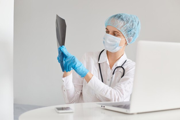 Dottoressa concentrata che indossa berretto medico, maschera chirurgica, guanti di gomma e camice, utilizzando notebook per lavorare, fare videoconferenza con i colleghi, mostrare raggi x.