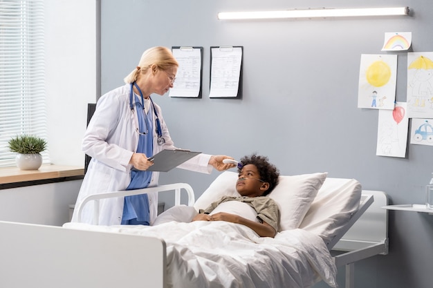 Dottoressa in camice bianco che tiene la tessera sanitaria e parla con un ragazzino che giace a letto nel reparto ospedaliero
