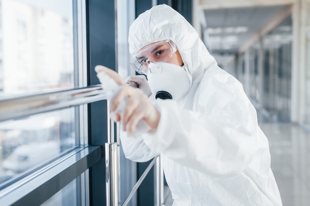 Dottoressa in camice da laboratorio, occhiali difensivi e maschera in piedi al chiuso con spray antibatterico