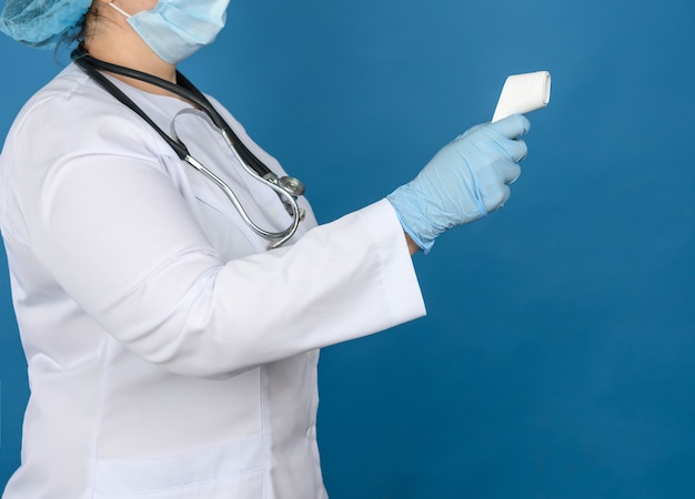 Dottoressa in camice medico bianco, guanti in lattice blu che tengono un termometro elettronico senza contatto in plastica, parete blu