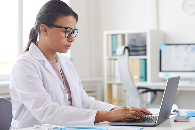 Dottoressa in occhiali e camice bianco seduto al tavolo e concentrandosi sul suo lavoro sul computer portatile