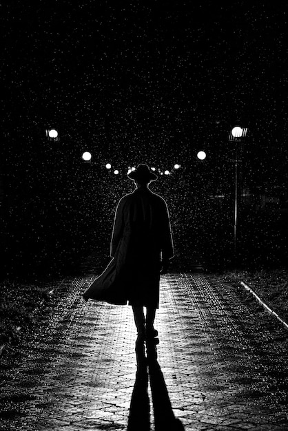 Drammatica silhouette di un uomo con cappello e impermeabile che cammina per la città di notte sotto la pioggia in stile Noir retrò