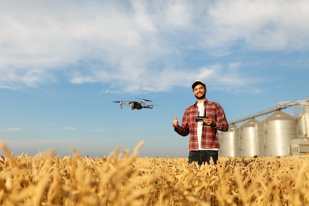 Drone hovers in front of farmer with remote controller in hands near grain elevator Quadcopter flies near pilot Agronomist taking aerial photos and videos in a wheat field