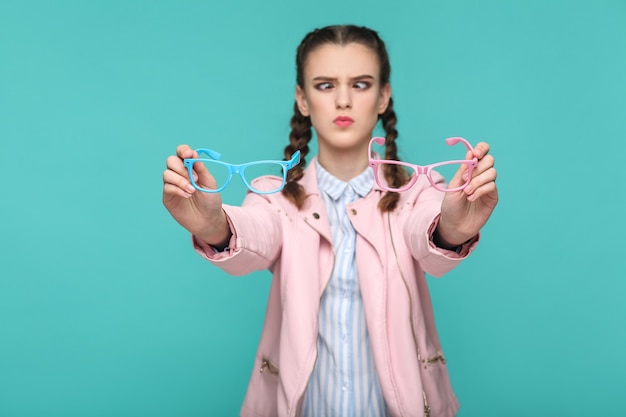 Dubbiosa ragazza divertente in stile casual o hipster, acconciatura a codino, in piedi, con in mano occhiali blu e rosa e guardando la telecamera con occhio incrociato, studio al coperto, isolato su sfondo verde