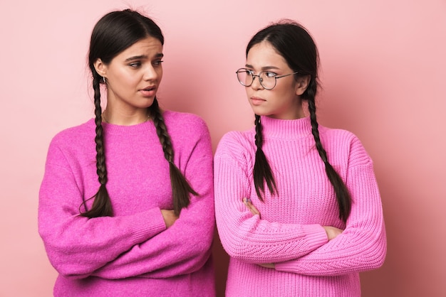 due adolescenti arrabbiati con le trecce in abiti casual in piedi con le braccia incrociate isolate su un muro rosa