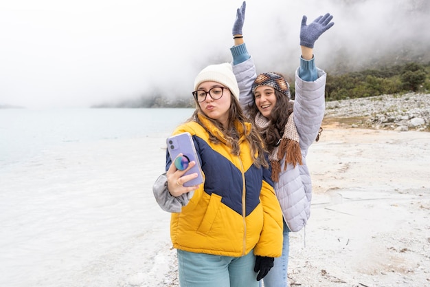 Due adolescenti che gesticolano in modo divertente mentre si fanno un selfie davanti a una laguna alpina