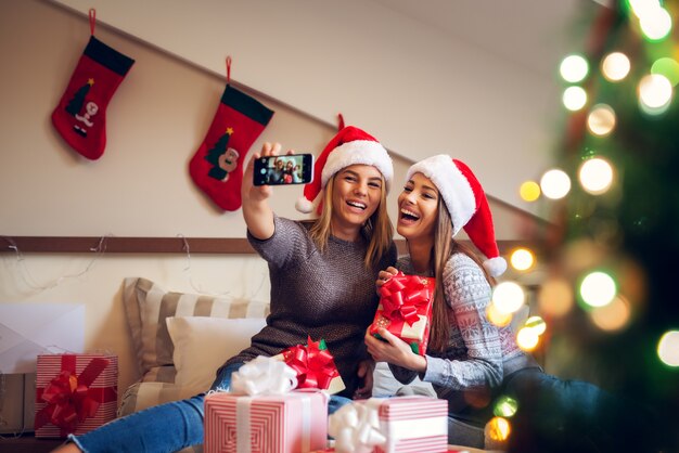 Due adorabili amiche con il cappello di Babbo Natale seduti sul letto per le vacanze di Natale una accanto all'altra e si fanno un selfie mentre ridevano di lode.