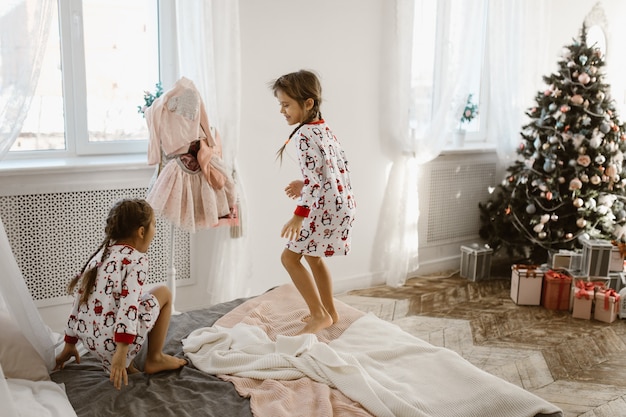 Due affascinanti bambine in pigiama si divertono a saltare su un letto in un'accogliente camera da letto illuminata dal sole con albero di Capodanno e regali. .