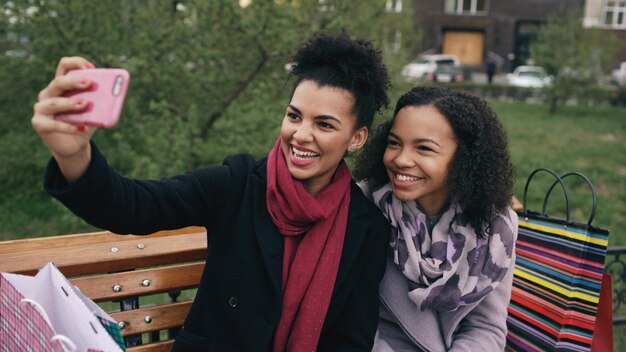 Due allegre donne di razza mista con borse della spesa che parlano in videochiamata con smartpone