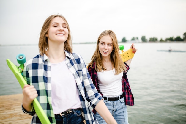 Due allegre ragazze pattinatrici felici in abito hipster che si divertono su un molo di legno durante le vacanze estive.