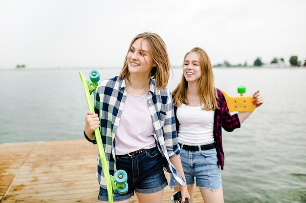 Due allegre ragazze pattinatrici felici in abito hipster che si divertono su un molo di legno durante le vacanze estive.